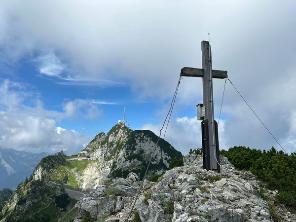 Ein Gipfelkreuz steht auf einem felsigen Berg mit einer malerischen Aussicht auf eine Gebirgskette und eine Senderanlage im Hintergrund.