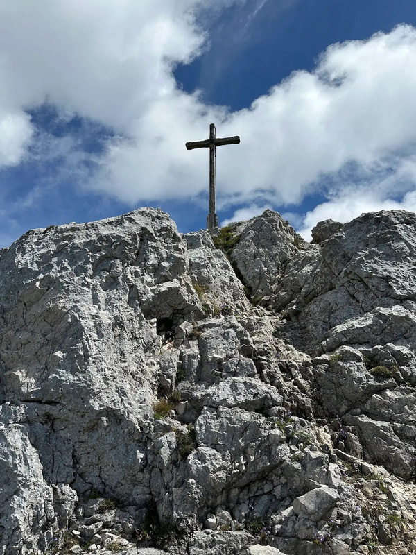 Ein Gipfelkreuz steht auf einem felsigen Berggipfel unter einem wolkigen Himmel.