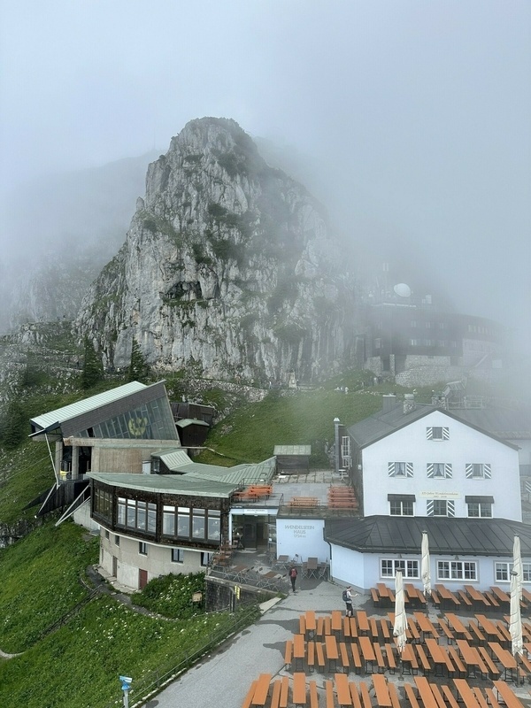Neblige Bergszene mit Gebäuden, Felsgipfel und leeren Bänken im Vordergrund.