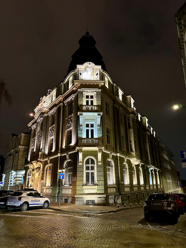 Ein beleuchtetes historisches Gebäude steht an einer Straßenecke in der Nacht, flankiert von geparkten Autos.