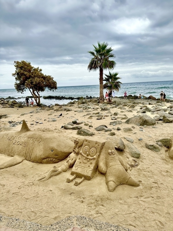 Ein Sandkunstwerk zeigt einen Hai, SpongeBob Schwammkopf und Patrick Star auf dem Strand in Maspalomas mit Palmen und bewölktem Himmel im Hintergrund.