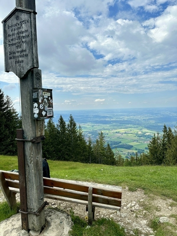 Eine Holzbank und ein Wegweiser stehen auf einem grasigen Gipfel, mit Blick auf Felder und Wälder unter teils bewölktem Himmel.