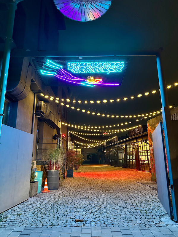 Ein gepflasterter Durchgang ist nachts von bunten Lichterketten und einem neonblauen Schild beleuchtet.