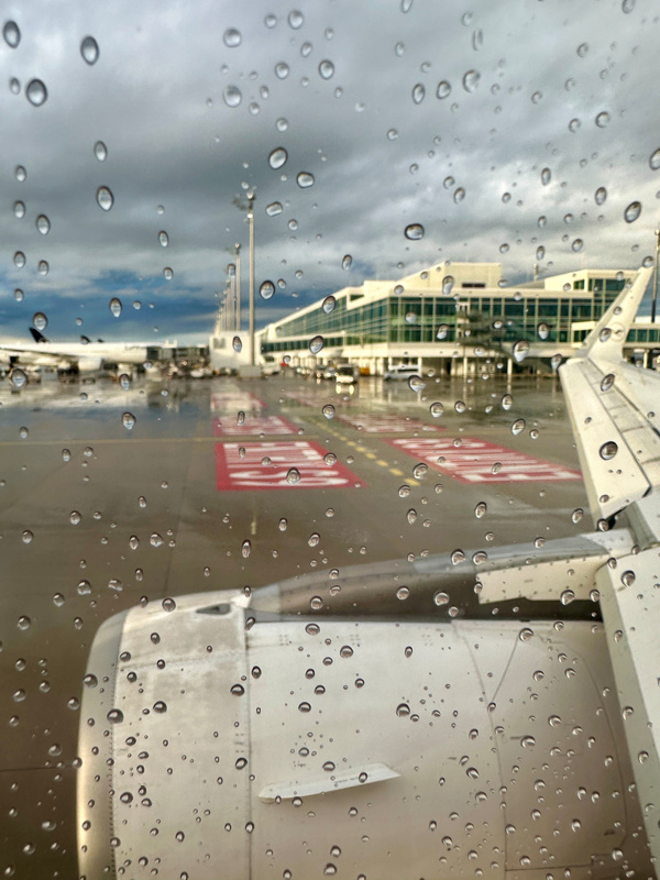 Ein Flugzeugflügel und ein Triebwerk sind durch ein regennasses Fenster auf einem Flughafen mit Terminalgebäuden und bewölktem Himmel sichtbar.