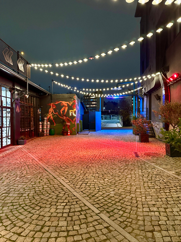 Eine gepflasterte Gasse ist mit Lichterketten beleuchtet und führt zu einem grafisch gestalteten Wandbild.