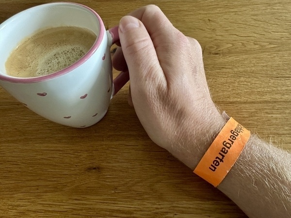 Eine Hand hält eine Tasse Kaffee, während sie ein orangefarbenes Armband mit der Aufschrift tagesgästen trägt.