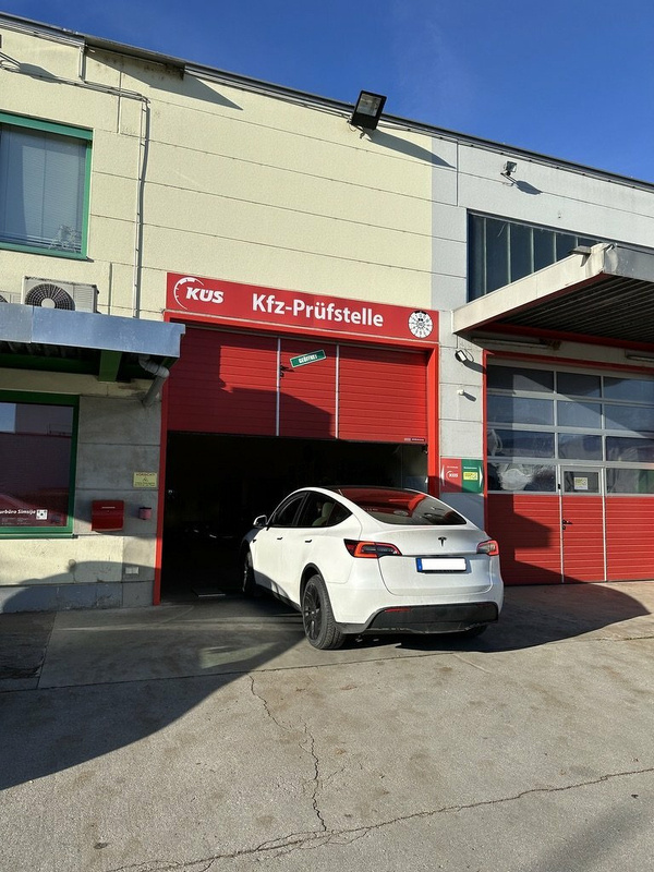 Ein weißes Tesla Model Y steht teilweise in einer Kfz-Prüfstelle unter einem roten Schild.