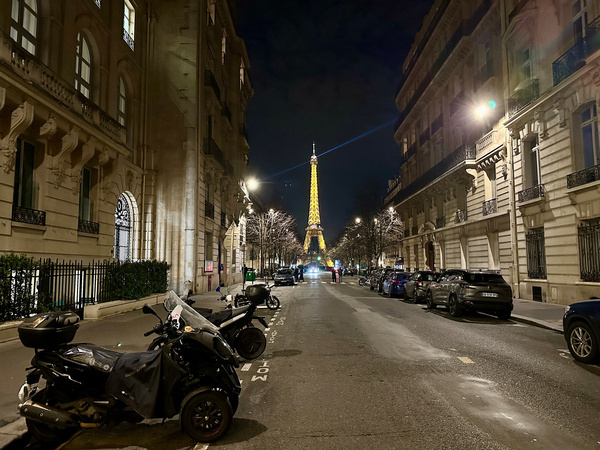 Straße in einer Stadt bei Nacht mit der beleuchteten Spitze des Eiffelturms im Hintergrund.