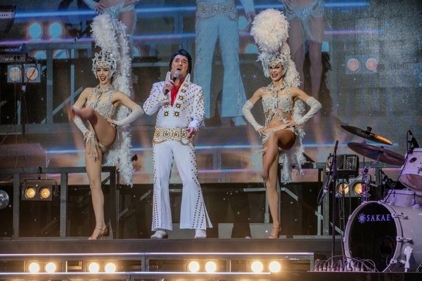 A performer dressed in a white jumpsuit reminiscent of Elvis Presley is flanked by two showgirls in elaborate costumes and feathered headdresses on stage.