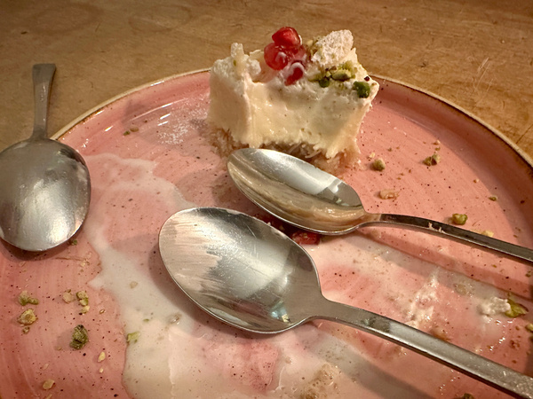 Ein halb gegessener Kuchen mit einigen Pistazienstücken und drei Löffeln auf einem rosa Teller.
