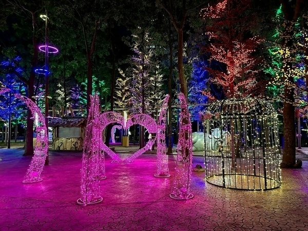 Ein lebendiges Nachtbild mit beleuchteten Bäumen, einer herzförmigen Lichtskulptur und einer käfigartigen Lichtinstallation auf farbigem Pflaster.