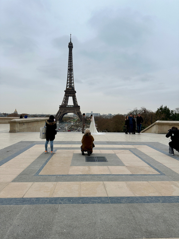 Ein Hochzeitspaar posiert vor dem Eiffelturm in Paris, während ein Fotograf und mehrere Personen sie fotografieren.