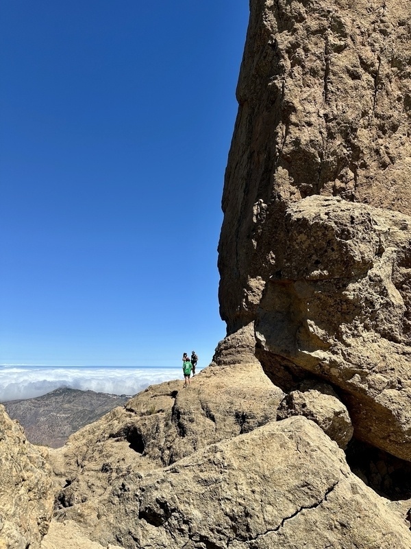 Zwei Personen wandern auf einem felsigen Pfad unter einem großen Felsvorsprung mit blauem Himmel im Hintergrund.