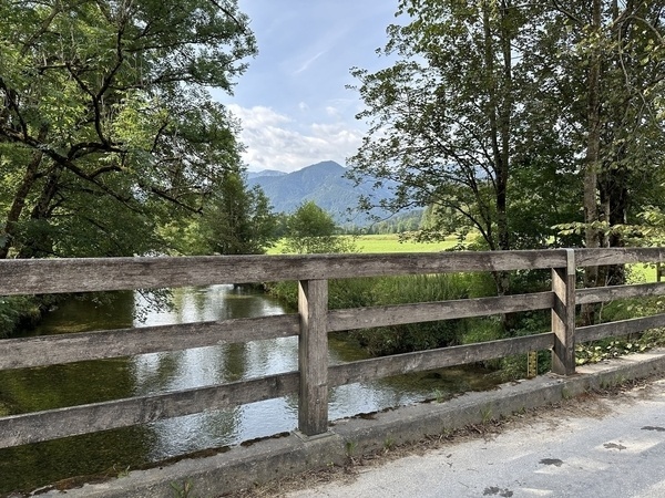 Eine Holzbrücke führt über einen friedlichen Bach, umgeben von üppigem Grün und fernen Bergen.