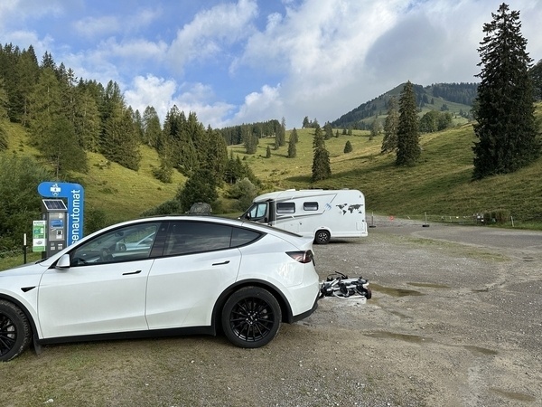 Ein weißes Auto mit Fahrradträger ist nahe einer Waschstation in einer bergigen Landschaft geparkt, im Hintergrund steht ein Campervan.