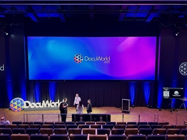 A stage setup for DocuWorld 2025 features a large screen displaying the event logo, with people discussing in front of it.
