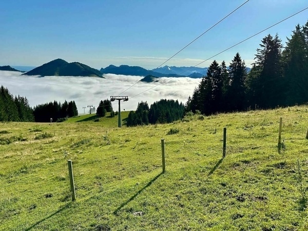 Ein malerisches Berglandschaftspanorama zeigt saftige Wiesen, umgeben von Wäldern und Wolken, mit einem Skilift im Vordergrund.