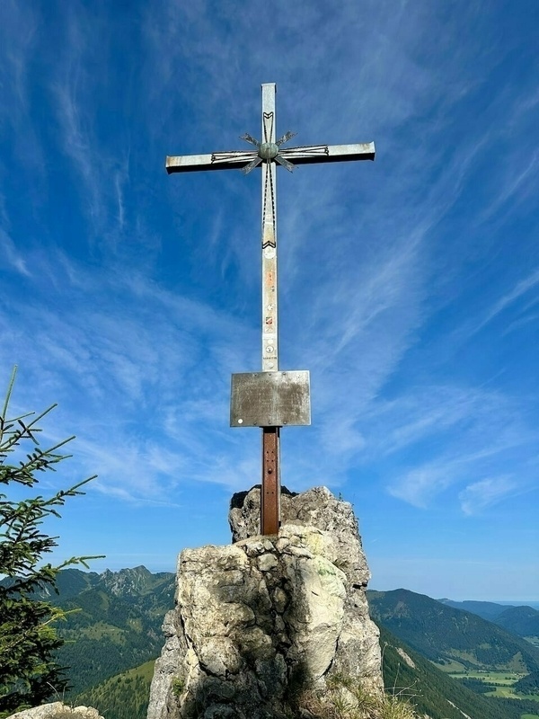 Ein Gipfelkreuz aus Metall steht auf einem Felsen mit einem blauen Himmel und Berglandschaft im Hintergrund.