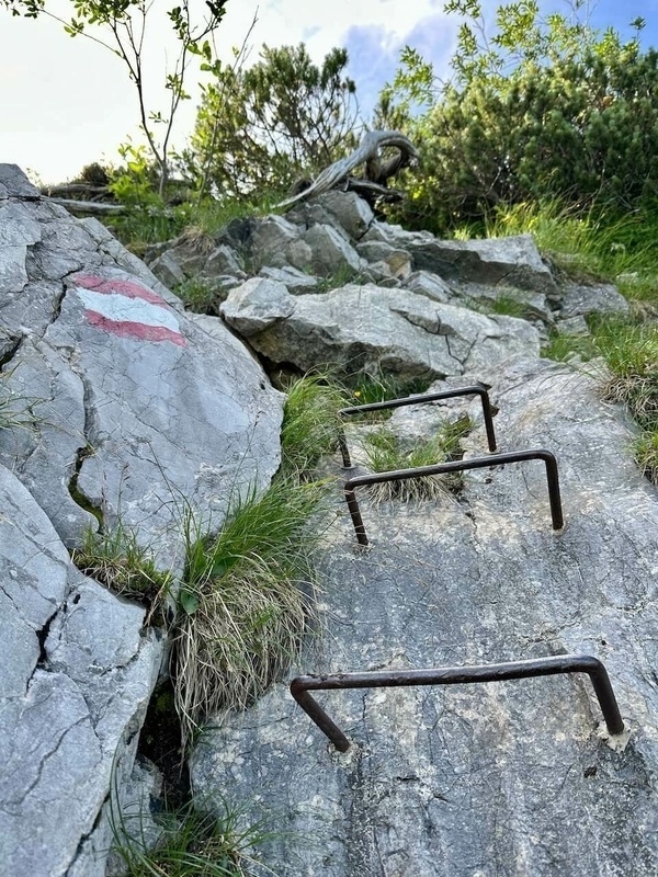 Metallstufen sind in einen felsigen Bergpfad eingelassen, daneben ist eine rot-weiße Markierung zu sehen.