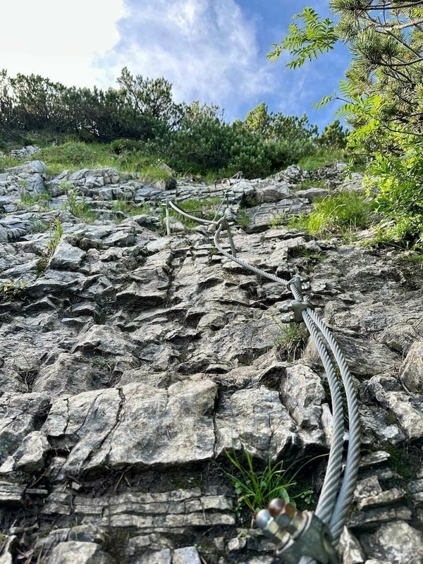 Ein Klettersteig führt einen steilen, felsigen Hang hinauf, gesichert durch ein Stahlseil.