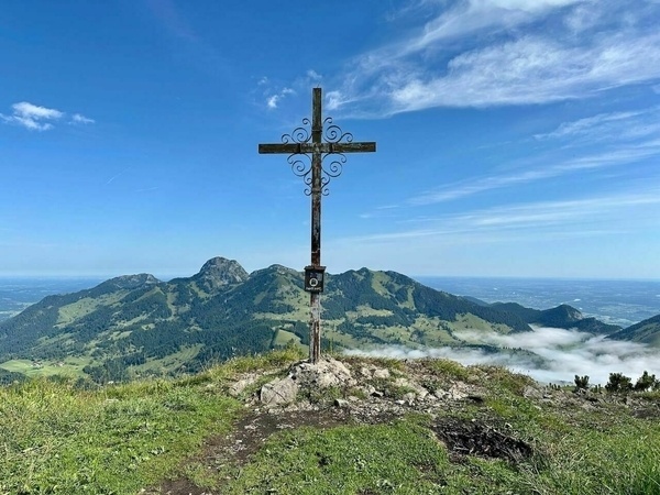 Ein Gipfelkreuz steht auf einem Berg mit einer weitreichenden Aussicht auf umliegende Berge und einen klaren Himmel.