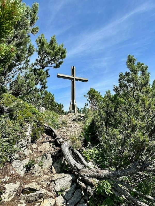 Ein Metalkreuz steht auf einem felsigen Pfad, umgeben von Kiefern und unter einem klaren blauen Himmel.