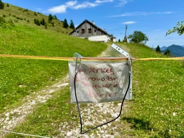 Ein mit handgeschriebenem Text versehenes Schild hängt an einem Kabel vor einer Wiese und einem Holzhaus in einer ländlichen Berglandschaft.