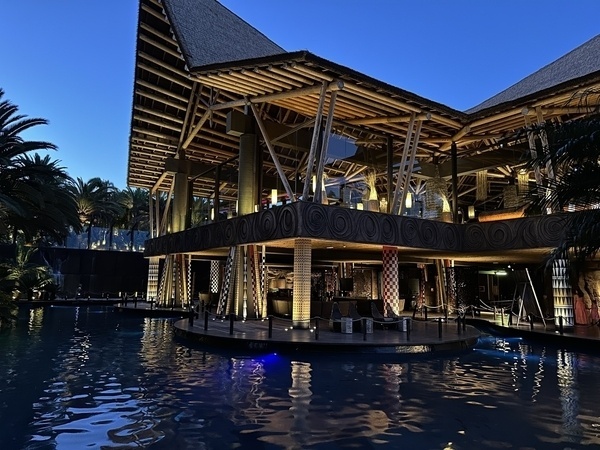 Ein stilvolles Gebäude mit Strohdach und hölzernen Elementen spiegelt sich in einem beleuchteten Pool bei Dämmerung wider.