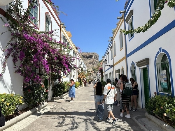 Eine sonnige Straßenszene mit blühenden Bougainvillea-Pflanzen und mehreren Menschen, die zwischen weiß gestrichenen Gebäuden mit farbigen Fensterrahmen spazieren.