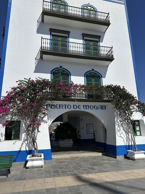 Ein weißes Gebäude mit blauen und grünen Details und einem Schild Puerto de Mogán wird von blühenden Pflanzen umrahmt.