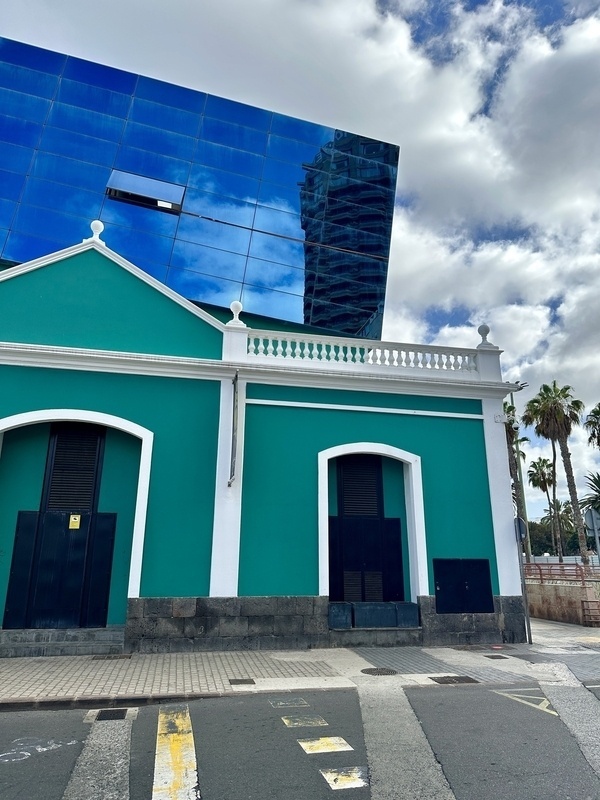 Ein türkis-blaues Gebäude mit weißen Akzenten steht vor einem modernen blauen Glasgebäude, während Wolken und Palmen den Hintergrund ergänzen.