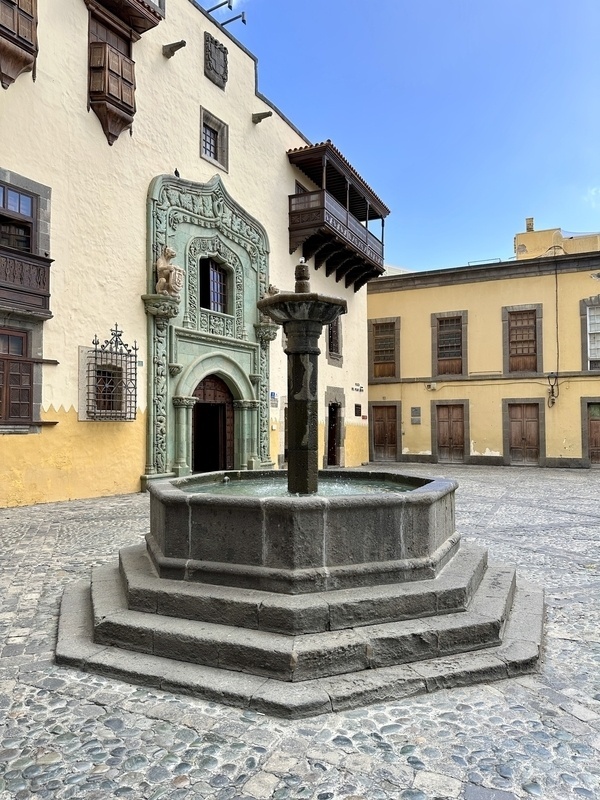 Ein historisches Gebäude mit einer kunstvollen Tür steht neben einem achteckigen Brunnen auf einem gepflasterten Platz.