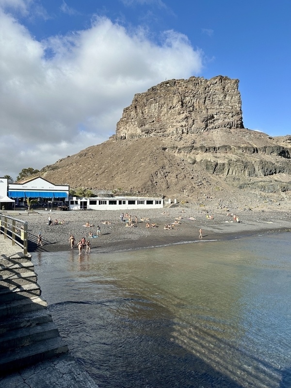 Ein felsiger Strand mit Menschen im Wasser und in der Sonne liegend, flankiert von einem großen, zerklüfteten Hügel und einem Gebäude am Ufer.