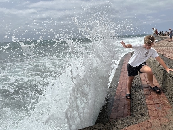 Ein Kind steht auf einer Promenade, während Wellen im Hintergrund gegen die Mauer schlagen.