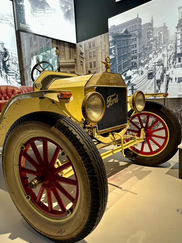 Ein historisches Ford-Automobil mit roten Speichenrädern steht in einem Ausstellungsraum vor alten Schwarz-Weiß-Fotos einer Stadt.