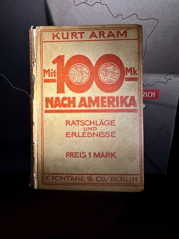 Ein Buch mit dem Titel Mit 100 Mk nach Amerika, geschrieben von Kurt Aram, liegt auf einer dunklen Oberfläche.