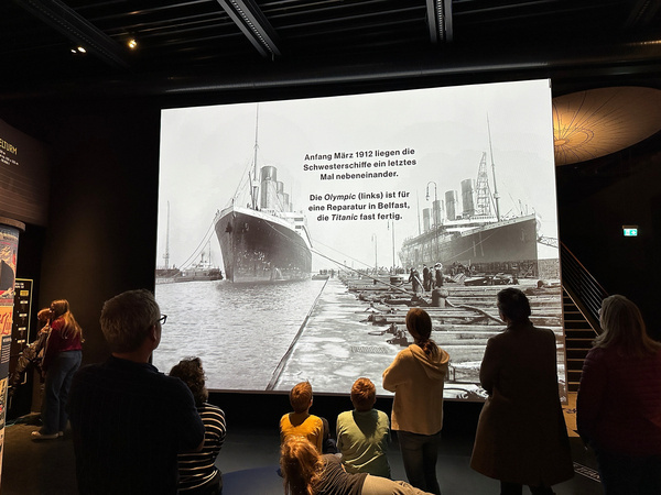 Menschen betrachten in einem Museum eine historische Schwarz-Weiß-Fotografie der Titanic und ihrem Schwesterschiff Olympic nebeneinander im Hafen.