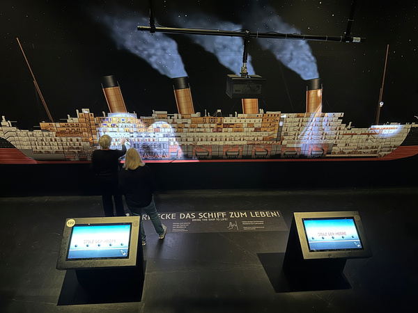 Zwei Personen betrachten ein illuminiertes Ausstellungsstück der Titanic mit interaktiven Bildschirmen.