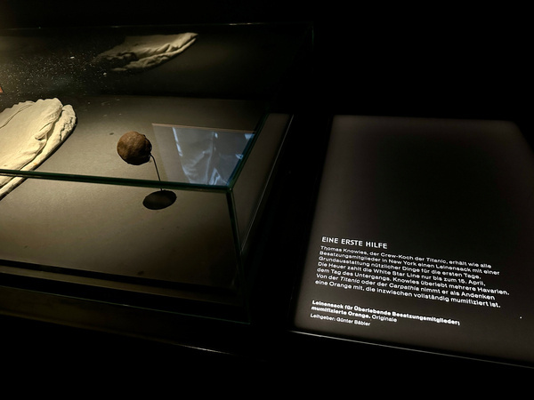 Eine Museumspräsentation zeigt eine mumifizierte Orange und ein Dokument mit Text über die Krise der Titanic, beschrieben in Deutsch.