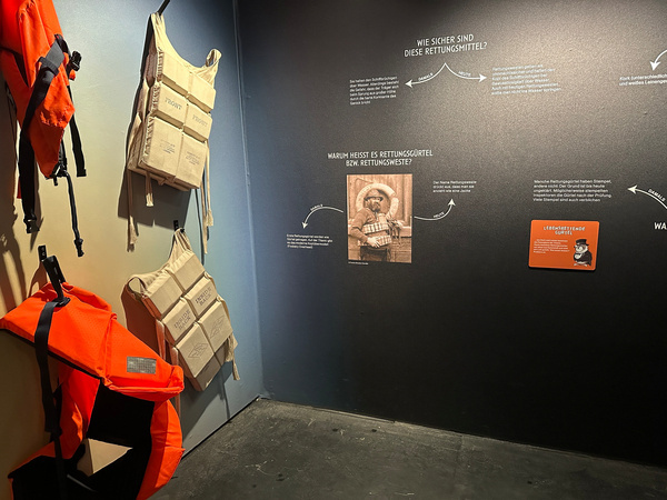 Ausgestellte Schwimmwesten an der Wand aufgehängt, begleitet von informativen Texttafeln zur Sicherheit von Rettungsmitteln.