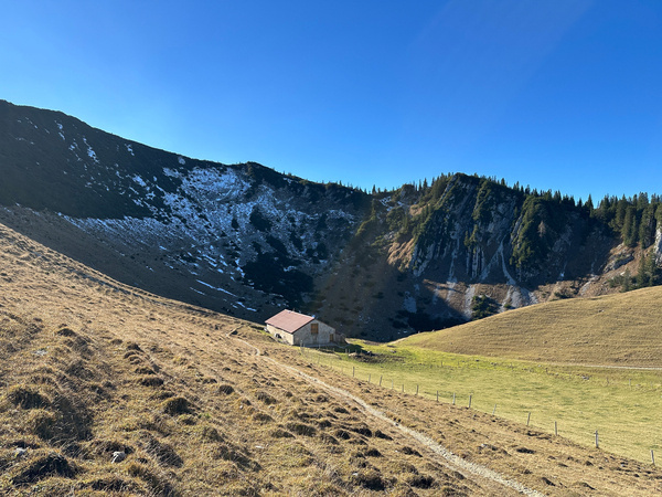 Die Jägerbauernalm vor dem Jägerkamp, einer grasigen Hügellandschaft mit schneebedeckten Bergen im Hintergrund unter einem klaren blauen Himmel.