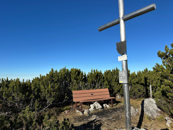 Das Gipfelkreuz der Benzingspitze steht neben einer Holzbank, umgeben von grünen Sträuchern unter einem strahlend blauen Himmel.