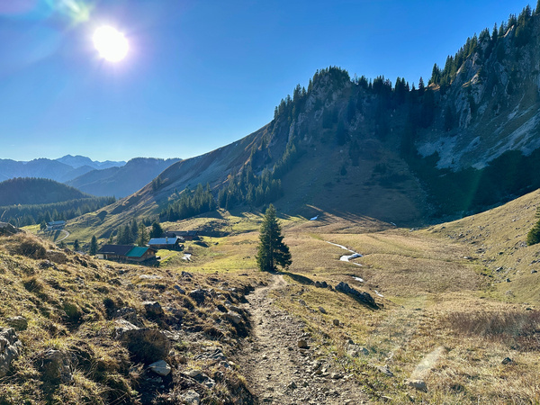 Eine malerische Berglandschaft mit Blick auf das Wilde Fräulein, zeigt einen sonnigen Tag mit einem klaren Weg, der durch ein alpines Tal mit einer Alm führt.