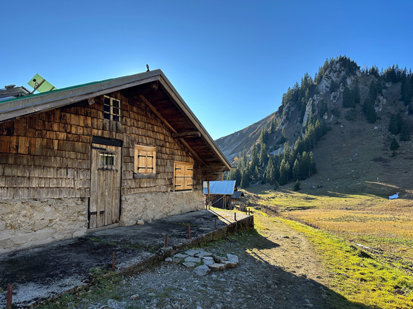 Eine rustikale Holzhütte steht in einer sonnigen Berglandschaft mit Blick auf das Wilde Fräulein und den Jägerkamp, mit einem bewaldeten Hügel im Hintergrund.