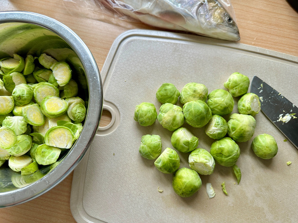 Rosenkohl und ein Messer liegen auf einem Schneidebrett, während mehr Rosenkohl in einer Metallschüssel liegt, und ein Fisch ist im Hintergrund zu sehen.