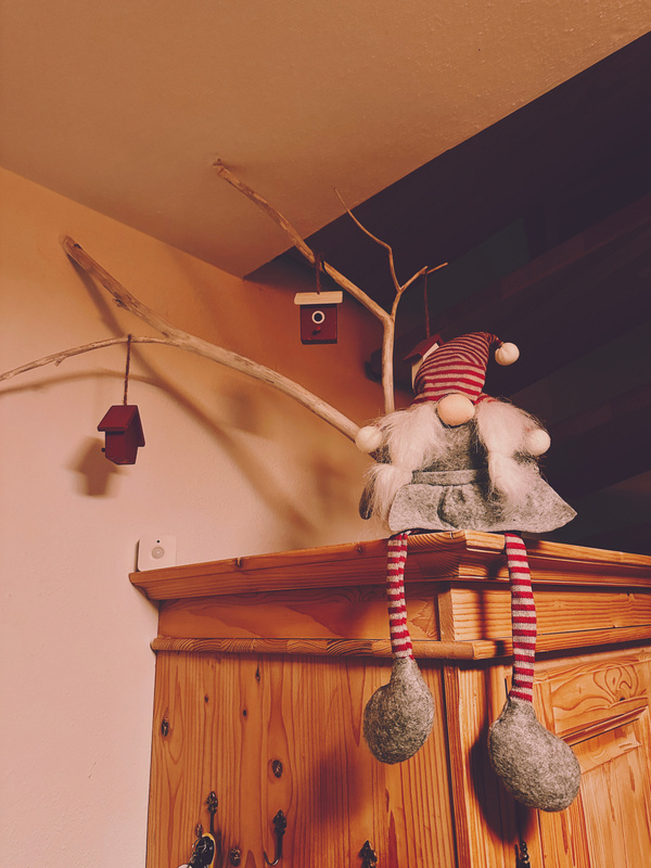 Eine dekorative Weihnachtswichtel mit gestreifter Mütze sitzt auf einem Schrank neben drei kleinen Vogelhäuschen an einem weißen Ast.