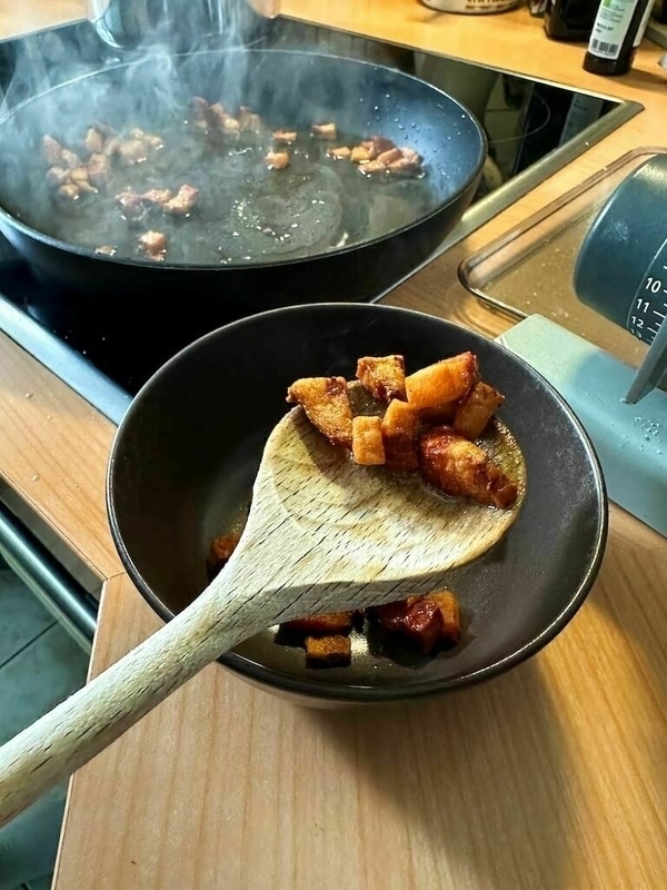 Gebratener Speck wird mit einem Holzlöffel aus einer Pfanne in eine weiße Schüssel verbracht.