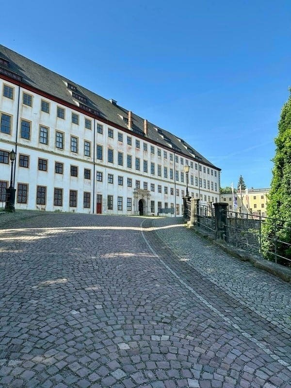 Das Schloss Friedenstein mit weißen Fassaden und zahlreichen Fenstern steht an einem gepflasterten Weg unter einem klaren blauen Himmel.