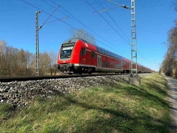 A red passenger train is traveling on a rail track through a scenic area with trees and a clear blue sky.