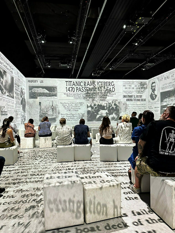 Menschen sitzen in einem Raum mit projiziertem Text und Bildern an den Wänden, die Berichte über die Titanic darstellen.
